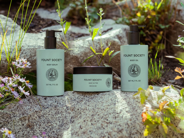 Three Fount Society skincare products, including body scrub, body butter, and body oil, displayed on natural rocks with surrounding greenery, showcasing a skincare bundle.