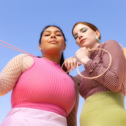 Two women posing confidently against a clear blue sky, wearing vibrant athletic wear from Bala's fitness destination, The Movement Store.