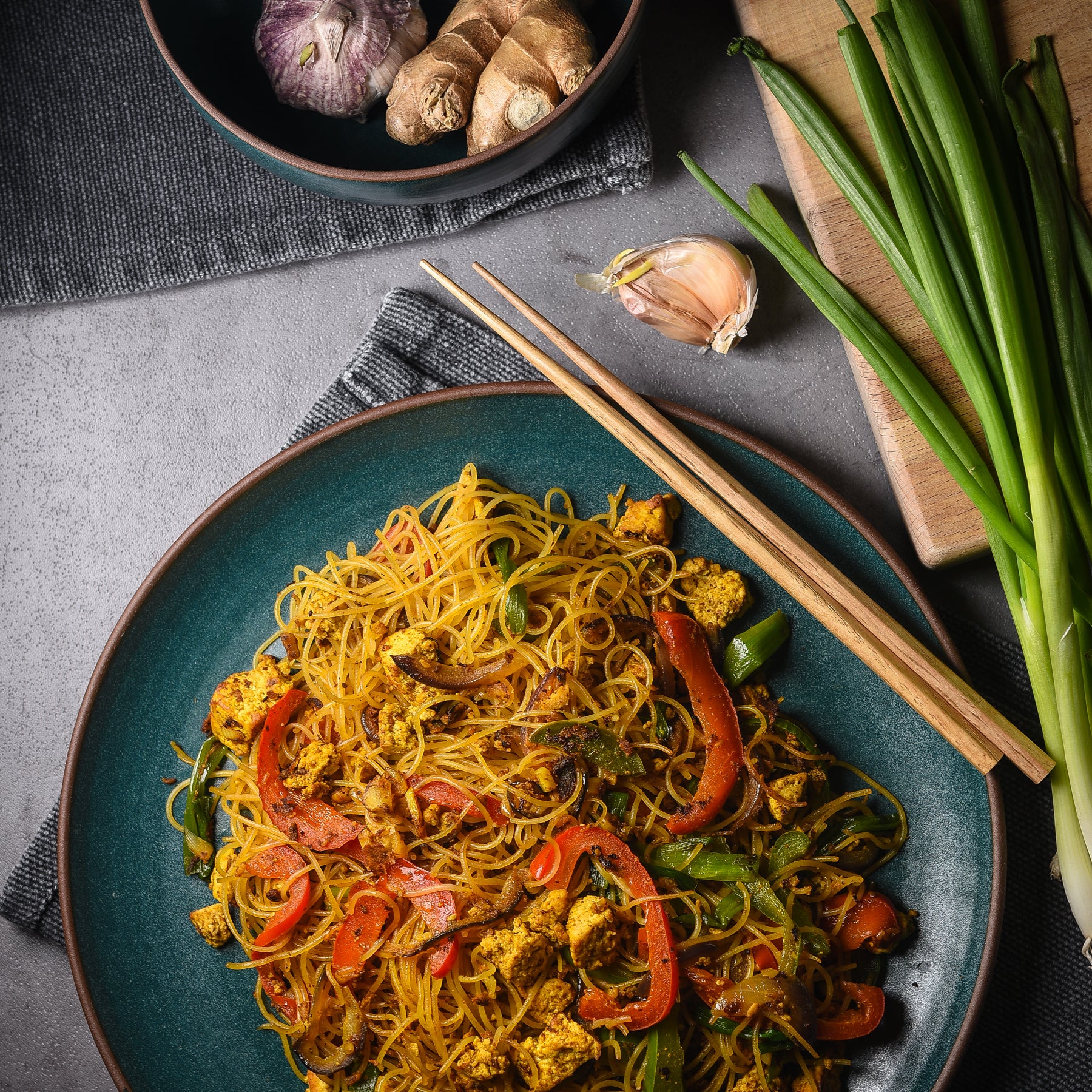 A plate of noodles with veggies surrounded by raw ingredients and chopsticks