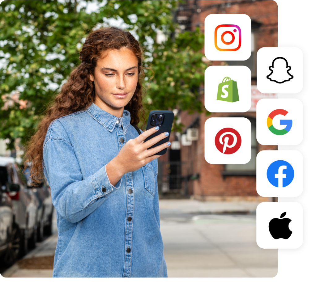 A white woman wearing a denim shirt stands on a tree-lined street and smiles as she checks her phone.