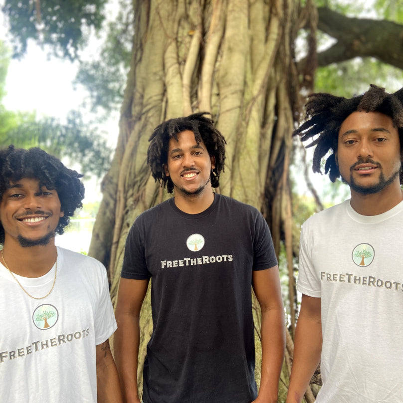 The three owners of FreeTheRoots stand together and smile in front of a large tree.