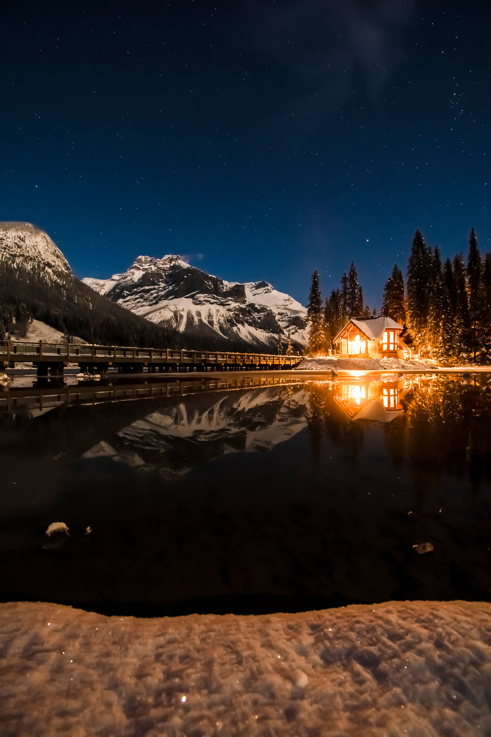 fire-lit house glows by a frosty lake against a starry sky